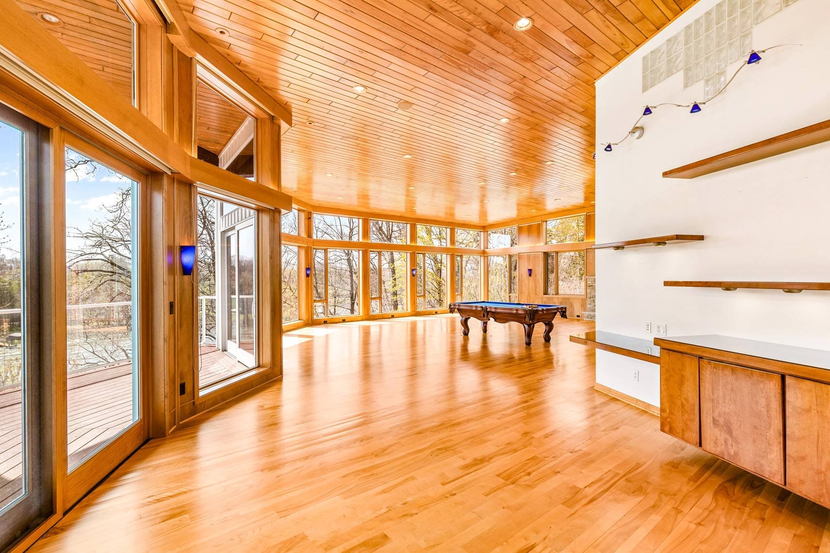 Residential property interior featuring a spacious living room with wooden floors and ceiling. Large windows provide natural light and offer views of the deck and surrounding trees. Room is furnished with a pool table and a shelving unit, ideal for family entertainment in a luxurious home setting.