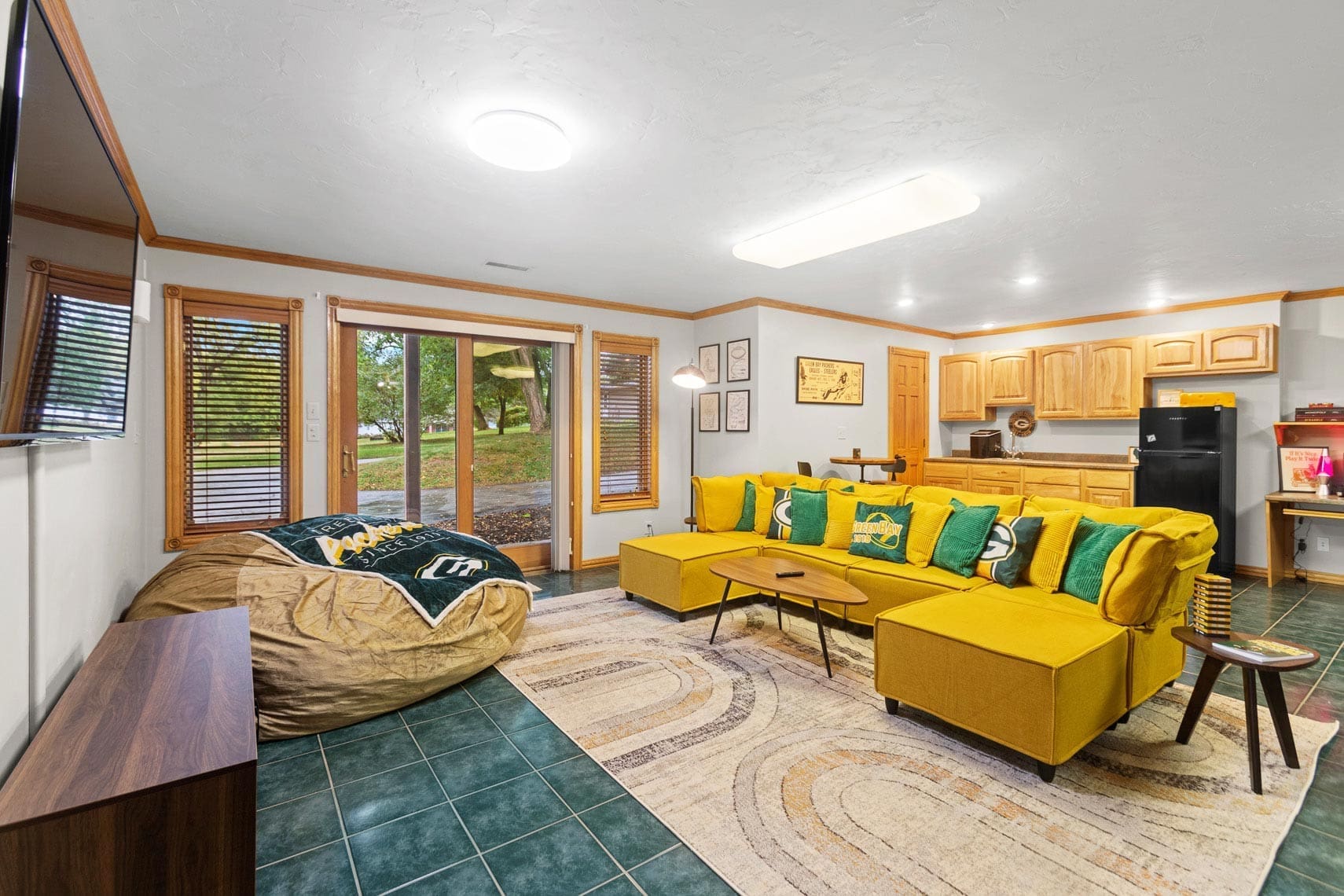 Residential living room featuring a yellow sectional sofa, bean bag, and wall-mounted TV with open-concept kitchen in the background. Large windows provide views of grassy outdoor area.