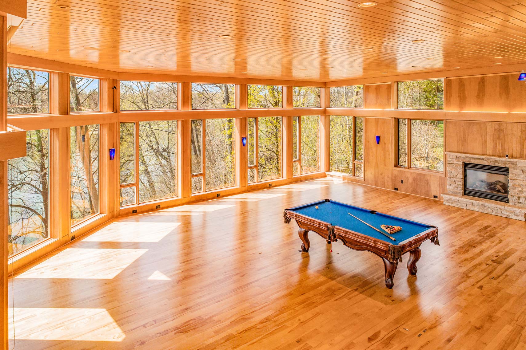 Photograph of a spacious residential property room featuring large windows, wooden flooring, light wood-paneled walls, a blue felt pool table in the center, and a stone fireplace on the side. Ideal for real estate listings showcasing entertainment spaces.