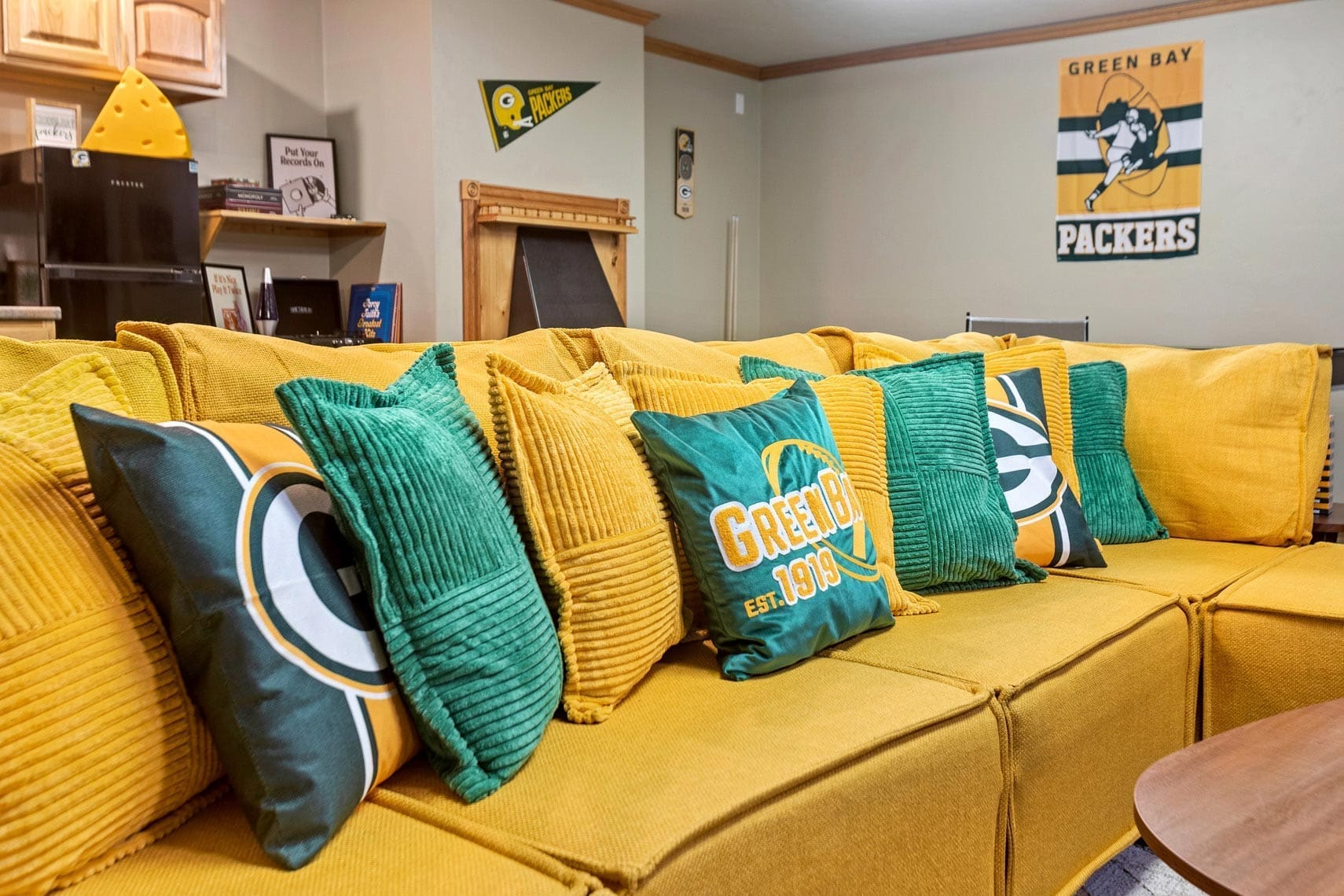 Residential real estate photography featuring a living room with a couch, yellow and green pillows, Green Bay Packers decor, and cheese-shaped hat for sports-themed home staging.