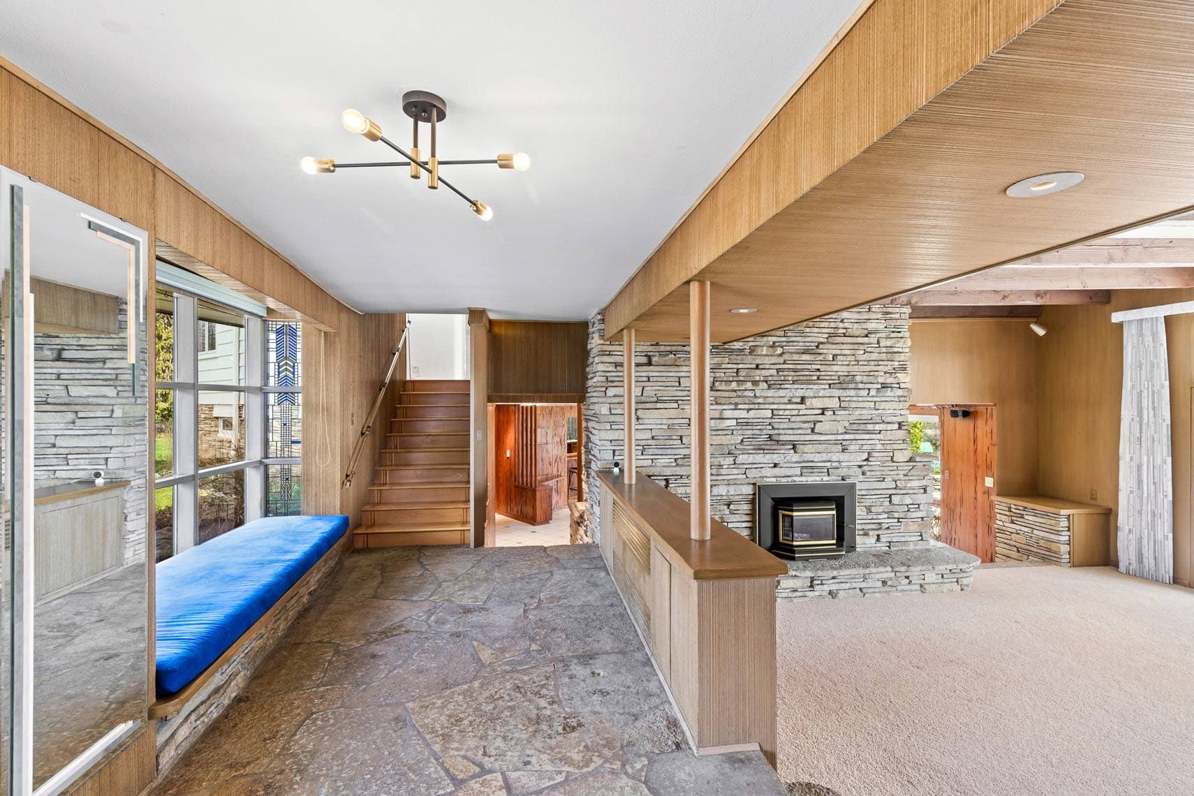 Residential property interior showcasing a room with stone flooring, wood paneling, and a stone fireplace. Stairs ascend from the hallway. Features a large window with a blue cushioned bench seat below it, ideal for natural light exposure. Ceiling light fixture complements the spaces design.