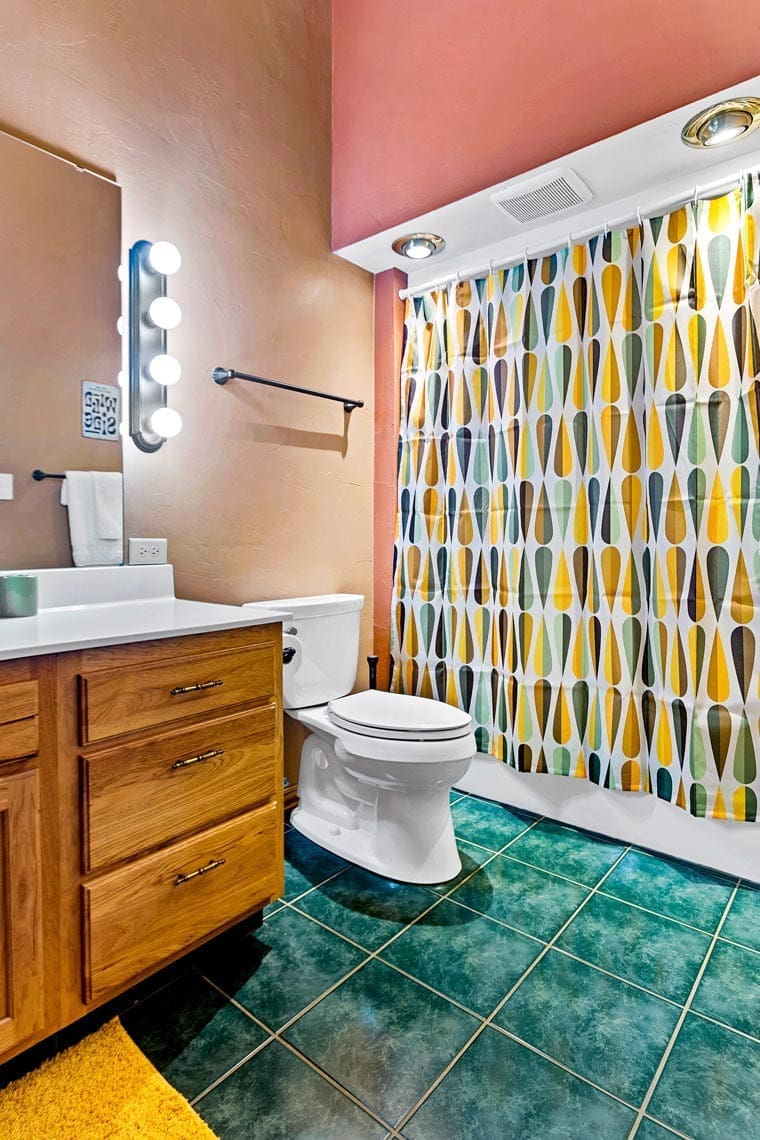 Residential bathroom featuring a wooden vanity, illuminated mirror, and white toilet; vibrant geometric shower curtain over the tub complements green tiled flooring. Perfect for real estate listings and vacation rental showcases.