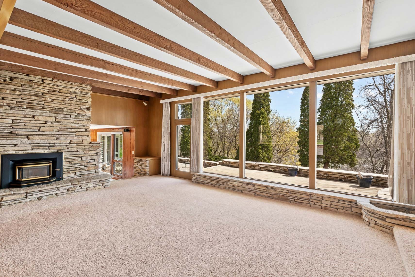 Residential real estate: Spacious living room featuring large windows, stone walls, a cozy fireplace, wooden ceiling beams, and an inviting outdoor view of lush trees.