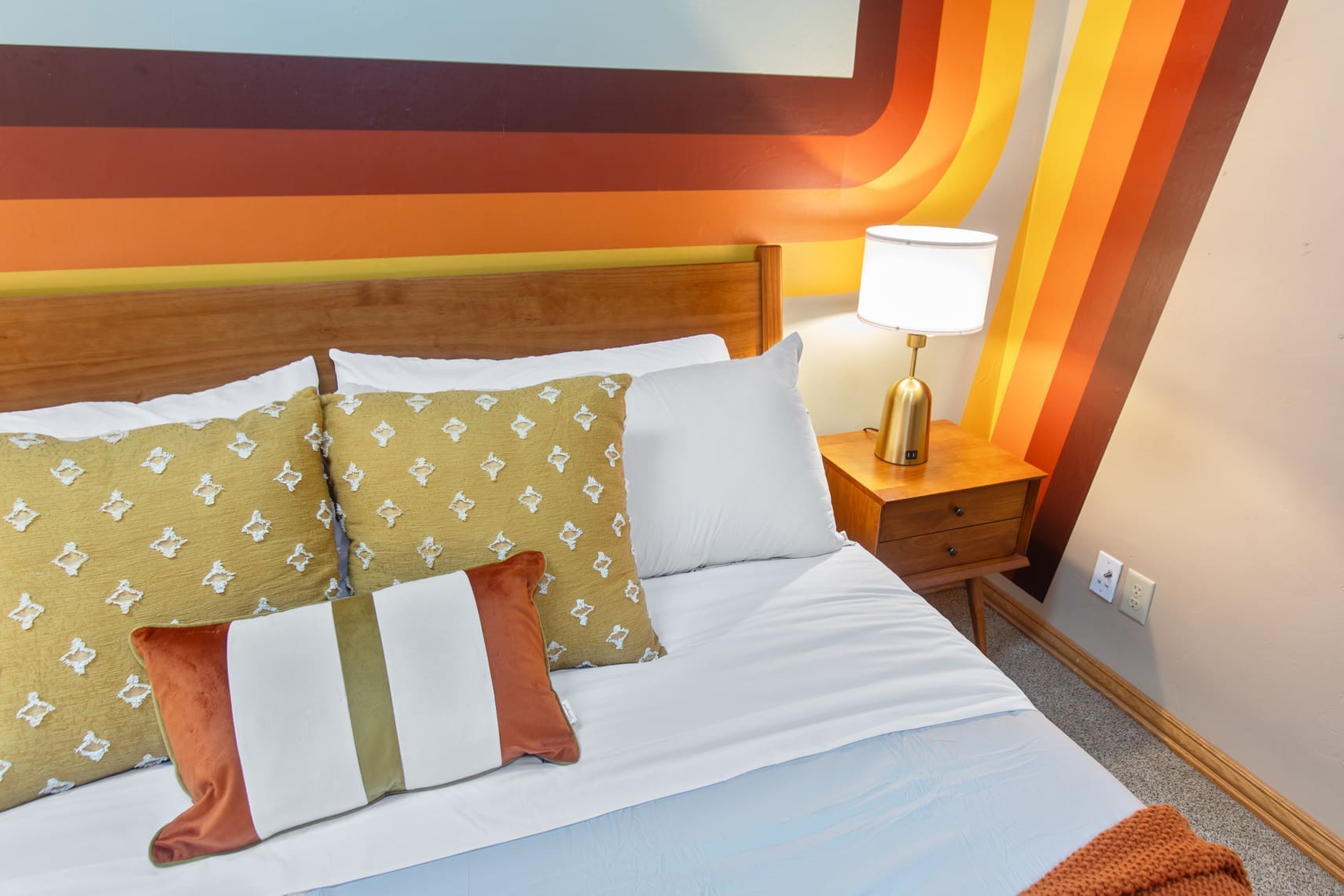 A professionally staged bedroom featuring a neatly made bed with yellow patterned pillows and a striped decorative pillow, ideal for real estate listings. A wooden lamp on the bedside table complements the retro-style striped wall in orange and brown tones, perfect for showcasing residential and vacation rental properties.