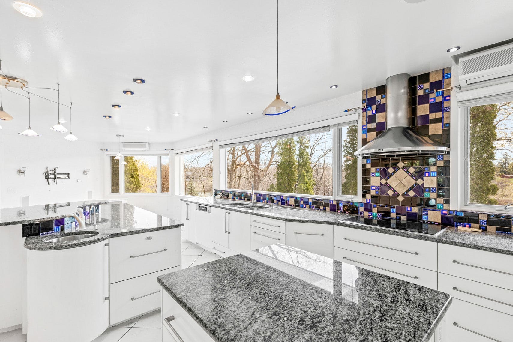 Spacious modern kitchen in a residential property, featuring glossy white cabinets, black granite countertops, and a colorful tiled backsplash. This real estate photo highlights the stainless steel oven hood and large windows offering tree views, perfect for commercial or vacation rental listings.