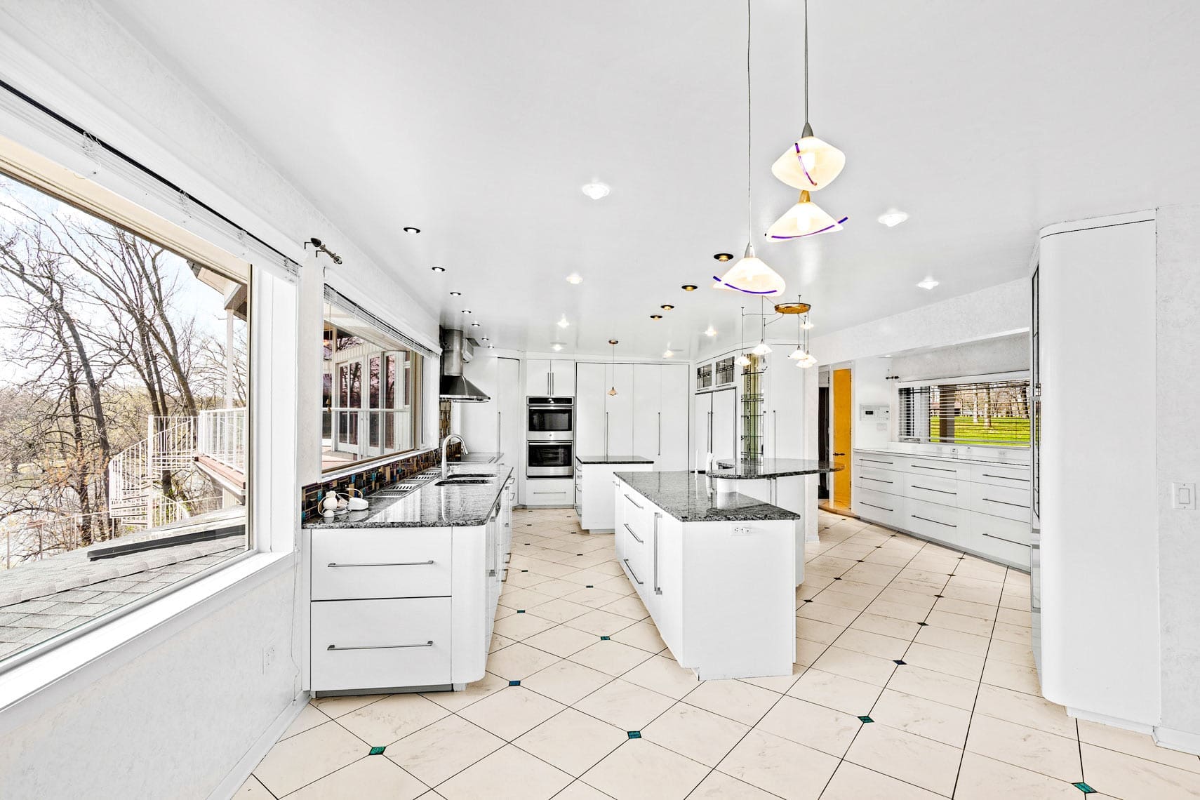 Expansive residential kitchen featuring large windows, contemporary cabinets, black countertops, and double center islands. Ideal for showcasing in real estate listings with its bright lighting and scenic outdoor views.
