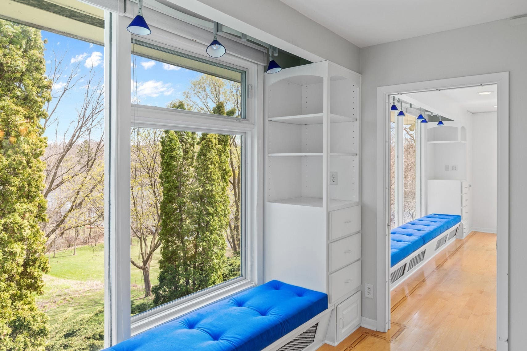 Residential property interior showcasing a bright room with a large window, white shelves, and a blue cushioned bench. Reflective mirror highlights ample storage space. Scenic view of trees and sky outside enhances the appeal.