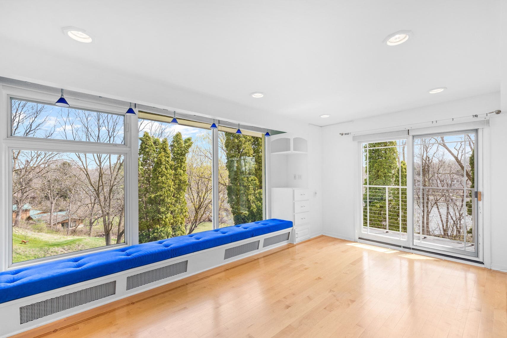 Bright residential space featuring large windows, a blue cushioned bench, and light wood flooring with scenic views of trees and distant homes. Glass doors on the right offer outdoor access. Overhead lighting enhances the commercial real estate presentation.
