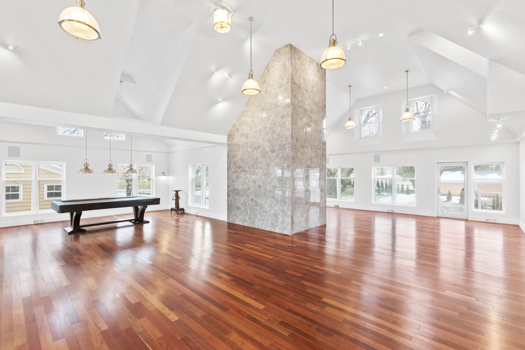 Elegant residential property showcasing a spacious interior with wooden floors, high ceilings, and multiple windows. Features include a central stone pillar, modern ceiling lights, and an upscale entertainment area with a pool table. Perfect for luxury real estate listings and commercial photography presentations.
