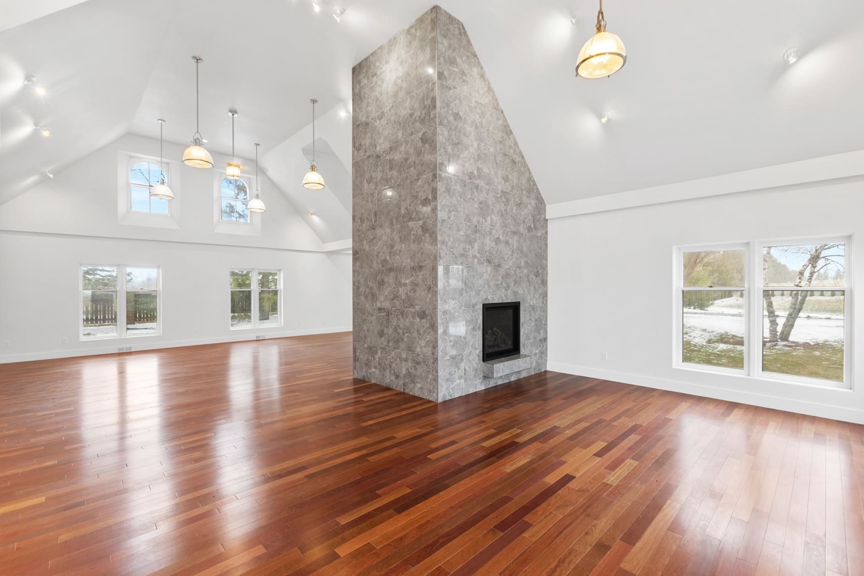 Spacious residential living room with high ceilings, elegant hanging lights, large windows for natural light, and polished wooden floors; features a central stone fireplace dividing the open-concept area.