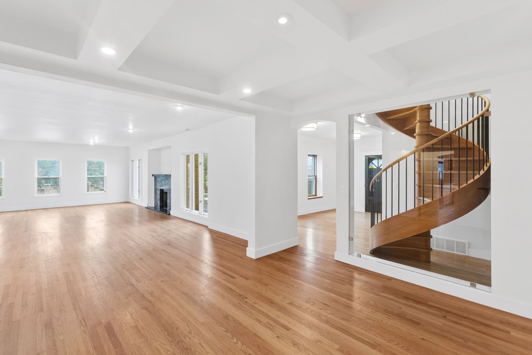 Open-concept residential real estate photo showcasing spacious living area with hardwood floors, bright white walls, expansive windows, and a striking wooden spiral staircase accented by ceiling lights.
