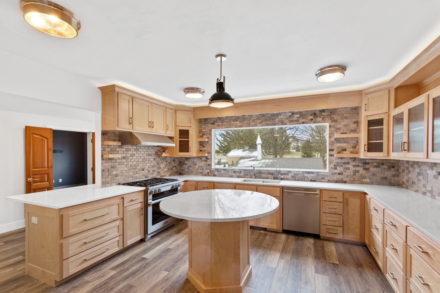 Real estate photography of a modern residential kitchen featuring wooden cabinets, a center island, stainless steel appliances, and a large window showcasing an outdoor view.