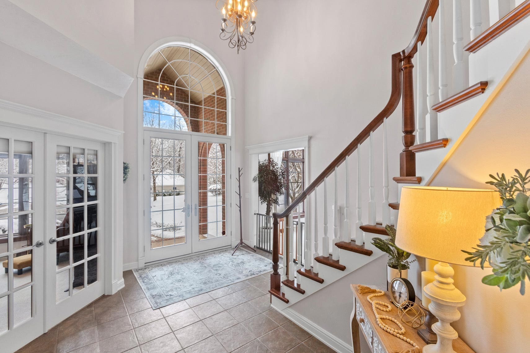 Spacious residential foyer with large arched window, elegant chandelier, and grand staircase in a real estate showcase. Glass doors on the left lead to a luxurious interior, while a lamp and decorative items add charm. Snow-covered landscape visible through the window enhances this propertys appeal for potential buyers.