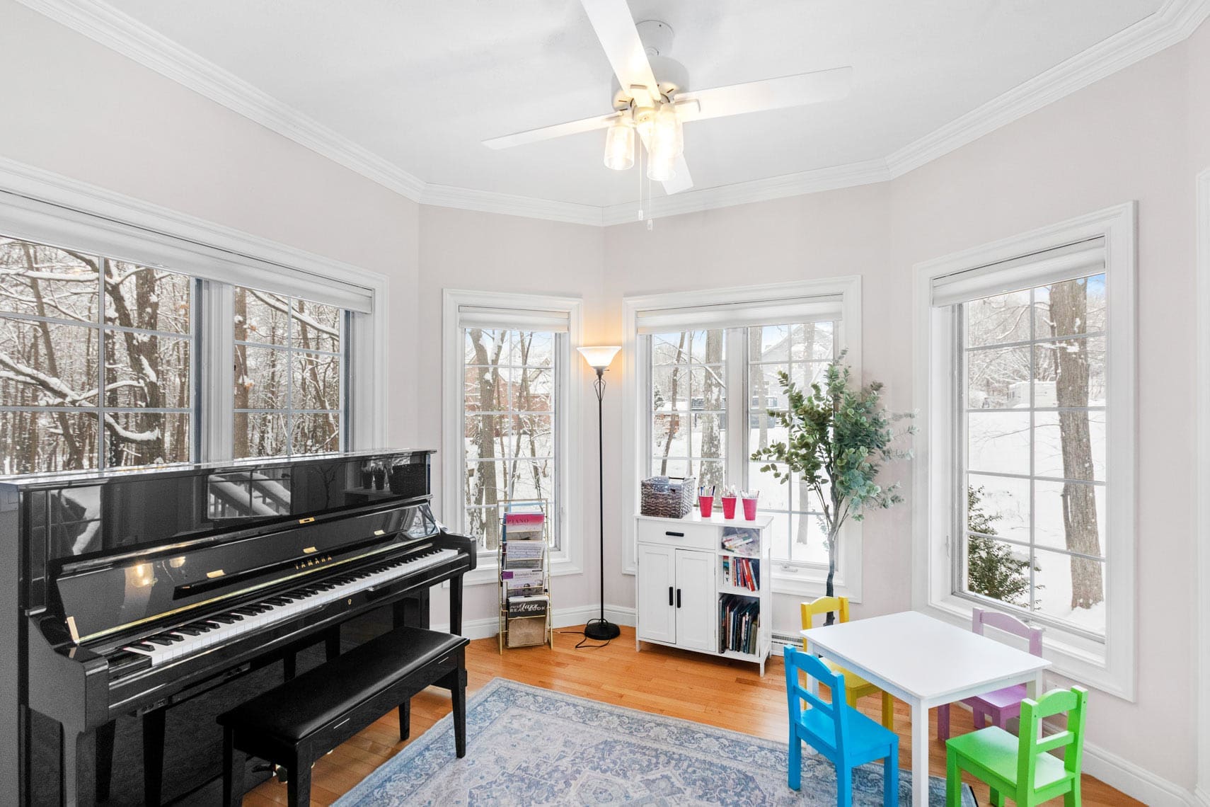 Residential property living room with large windows, a black piano, a white bookshelf with books, colorful chairs around a small table, and a ceiling fan. Snow-covered trees visible outside.
