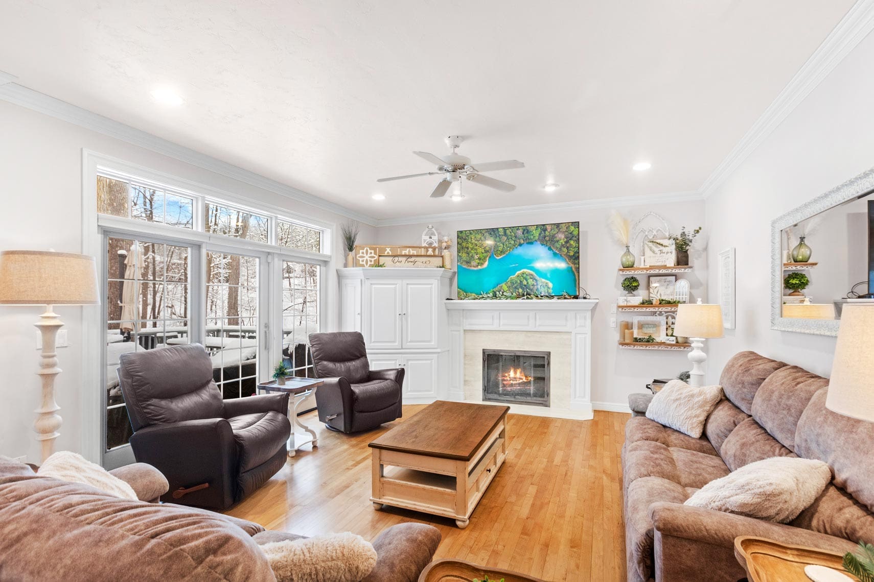 Residential real estate living room featuring brown sofas and armchairs, a coffee table, fireplace, wall-mounted TV, and large windows showcasing a tree-lined view. Thoughtful decor includes plants and shelves flanking the fireplace.