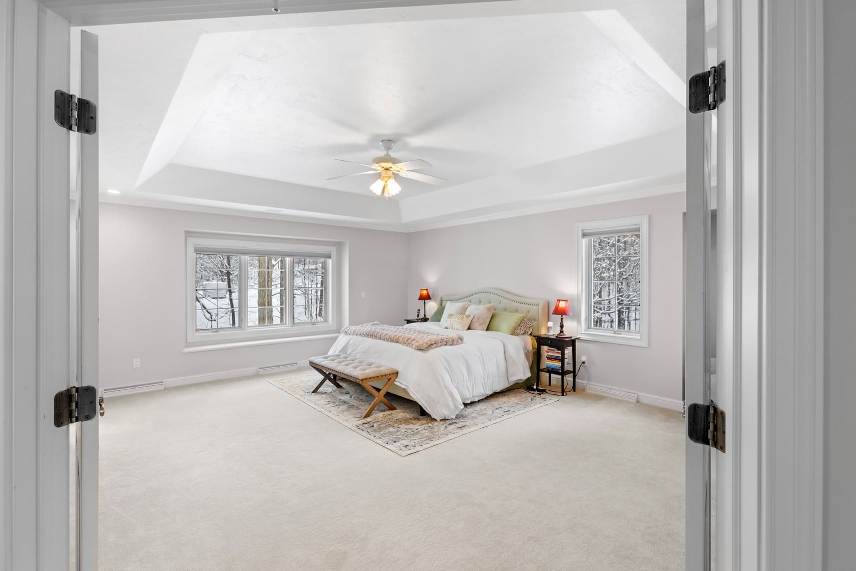 Residential real estate photography showcasing a bright and spacious bedroom with a large bed, two windows providing ample natural light, a ceiling fan for comfort, and bedside tables with stylish lamps. Features cream carpet and neutral walls, with double doors enhancing the welcoming atmosphere.