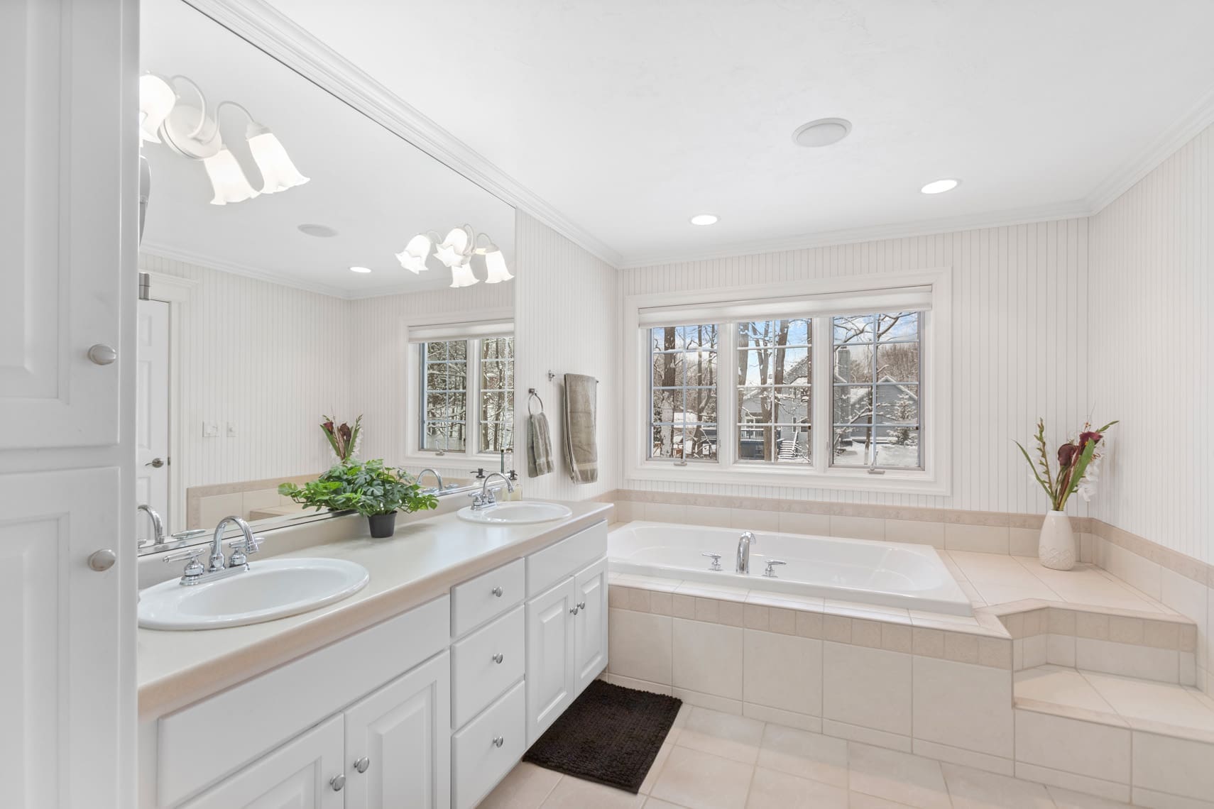 Bright residential bathroom featuring double sinks, a spacious bathtub with scenic window view, and white cabinetry. Beige tiles highlight this inviting space. Stylish elements such as a small plant and decorative vase enhance the commercial appeal of this real estate listing.
