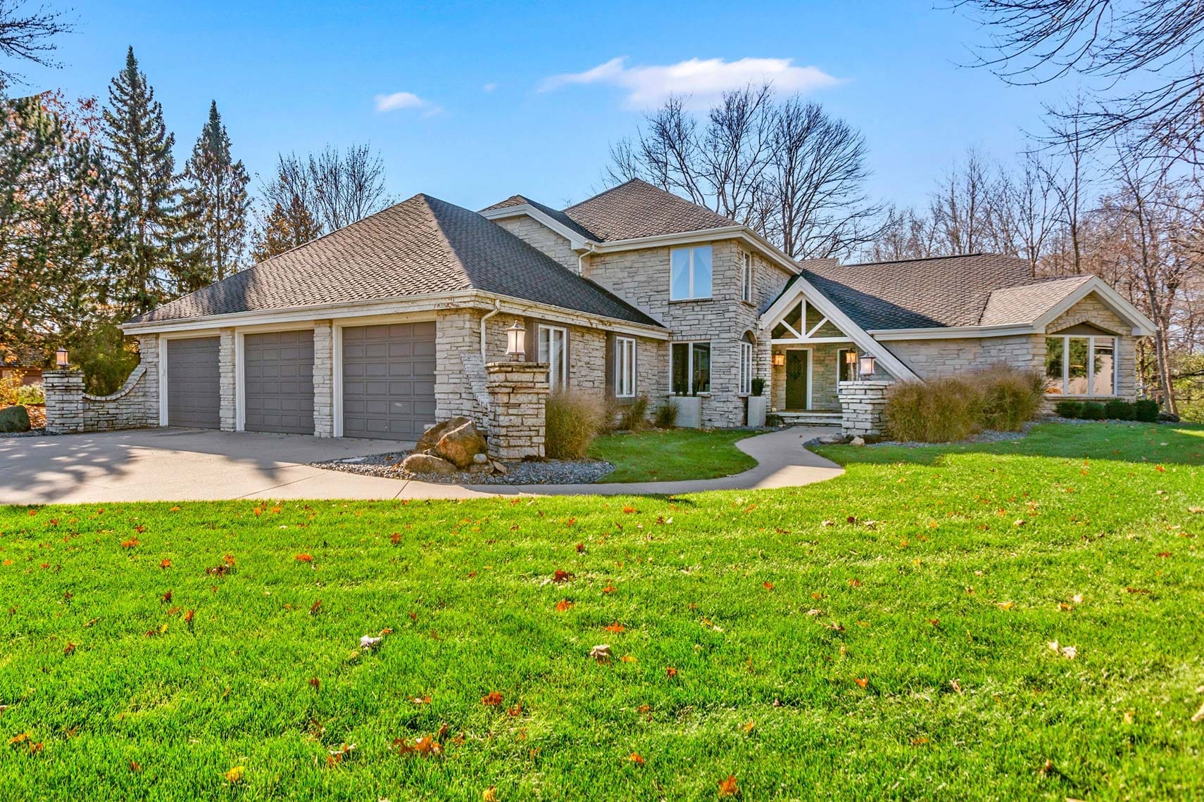 High-end residential property featuring a large stone house with multiple gabled roofs, a spacious three-car garage, and a meticulously manicured lawn. Sparse trees and clear blue sky enhance the luxurious real estate setting.
