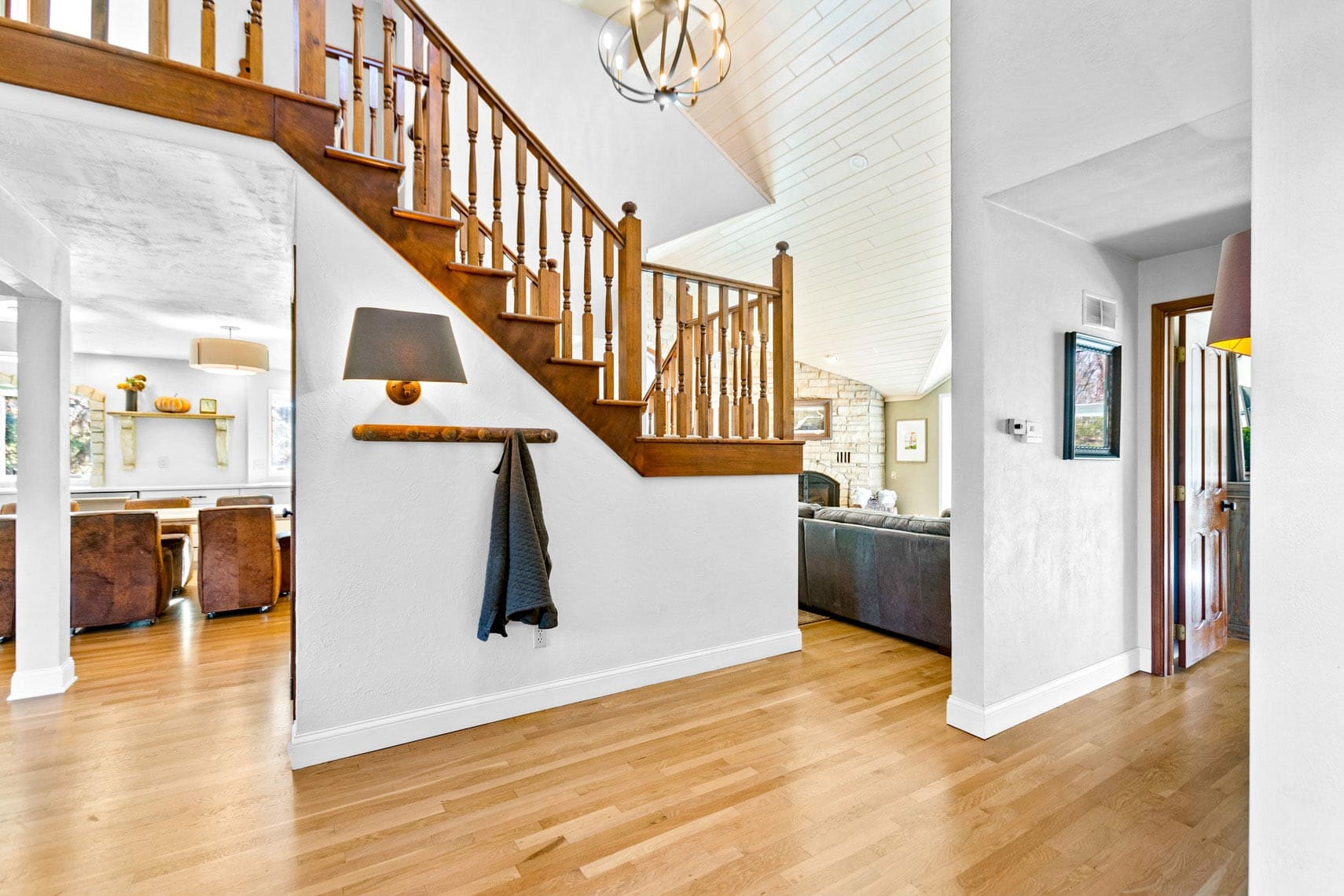 Interior of a spacious residential property featuring a wooden staircase, light hardwood floors, hanging lamp, and open living area. Ideal for showcasing real estate appeal with its seamless flow into the dining room accentuated by large windows.