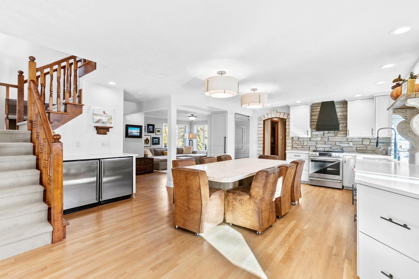 Residential real estate: spacious kitchen featuring wooden floors, modern appliances, stone backsplash, central island with leather chairs; open living area and staircase on the left.