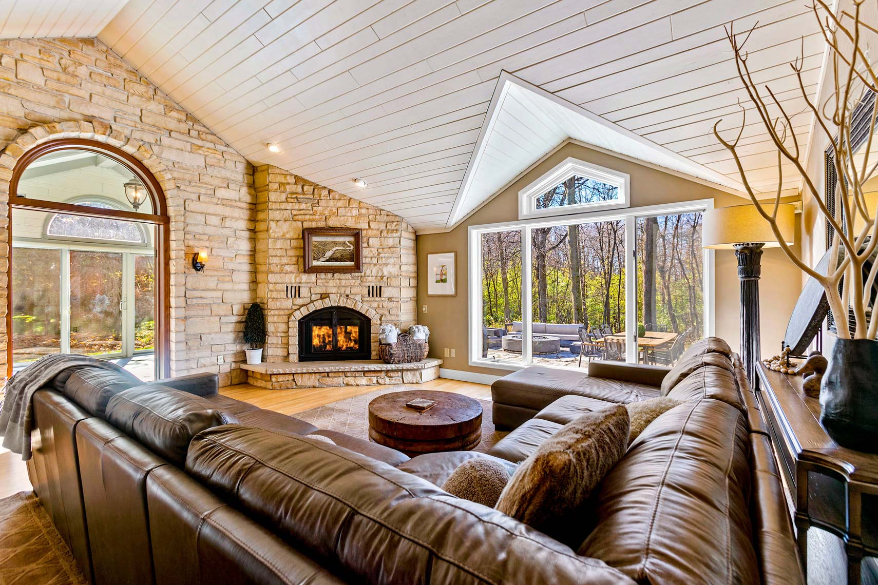 Residential real estate photograph showcasing a spacious living room with vaulted ceiling, large windows for ample natural light, brown sectional sofa, stone fireplace, and stunning outdoor view. Art-adorned walls and mounted light fixtures enhance the interior appeal.