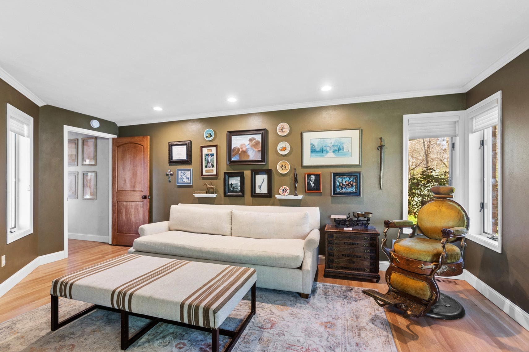 Real estate photo of a residential living room showcasing green walls, a white sofa, striped ottoman, vintage dentist chair, and a gallery wall of framed pictures. Features brown wooden door and two windows for natural light.
