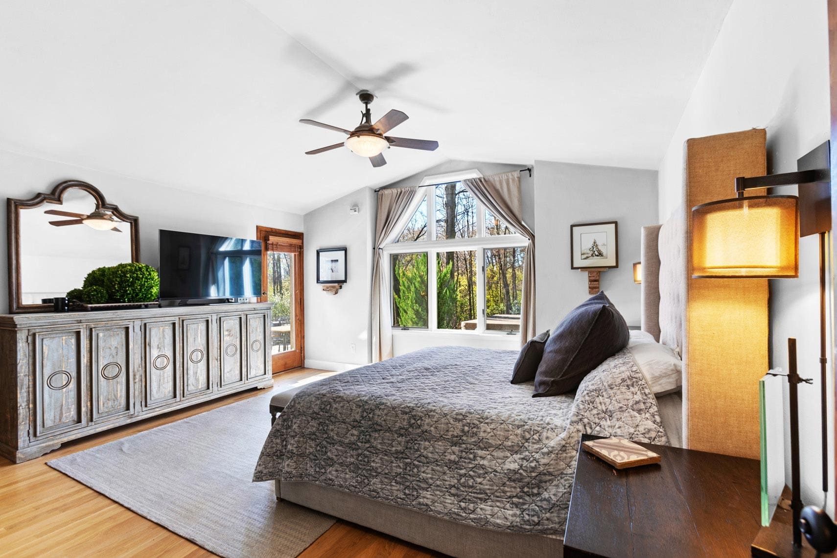 Bright bedroom in residential property with large bed, wooden floor, ceiling fan, wall-mounted TV, and dresser with mirror. Two windows offering natural light and picturesque greenery view. Ideal for real estate listings.