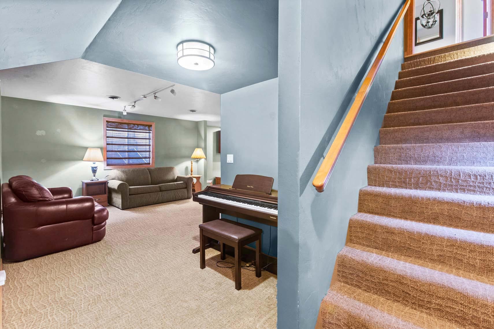 Residential basement interior showcasing carpeted stairs leading to a cozy living space with an electric piano and bench, leather armchair, couch, and ambient lamp. Green-painted walls adorn this inviting area for potential homeowners or renters, featuring a decorative framed picture above the stairs.