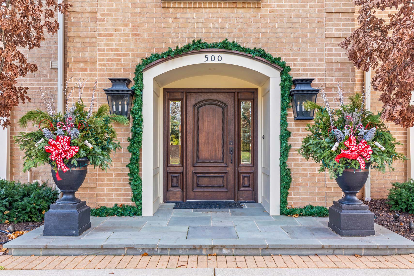 Residential property entrance featuring a wooden door with decorative arch, flanked by two urn planters with greenery and red bows, set in a brick building numbered 500.