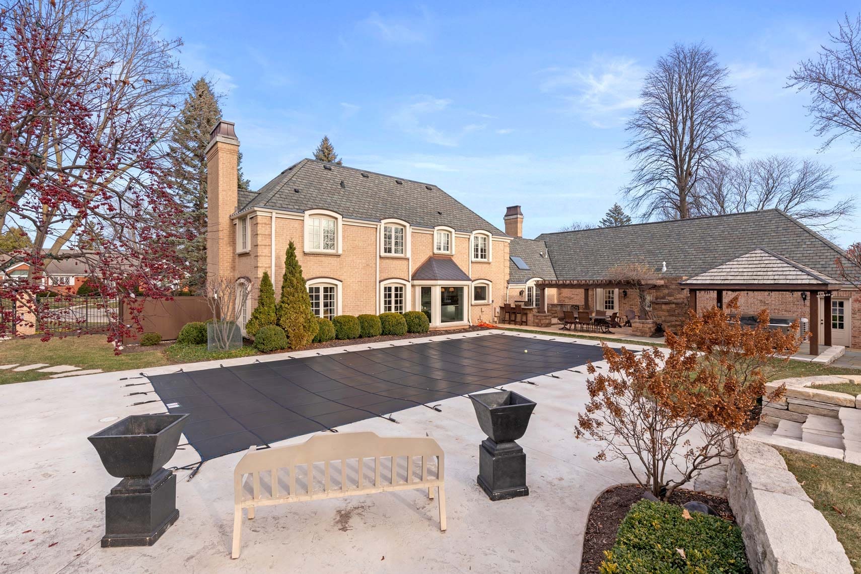 Expansive brick residential property featuring a covered rectangular pool, patio area, and nearby trees; perfect for real estate listings. Front wooden bench adds a cozy touch to this photographed home setting.