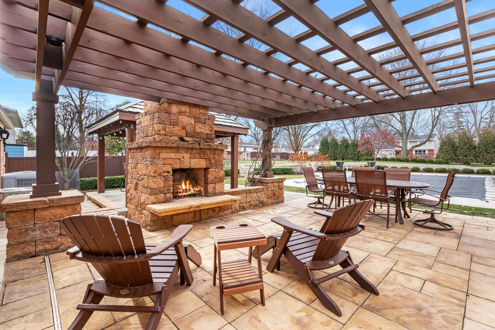 Commercial real estate photograph of an outdoor patio featuring a wooden pergola, stone fireplace, Adirondack chairs, small tables, dining set, and adjacent pool in a residential neighborhood with trees and houses visible in the background.