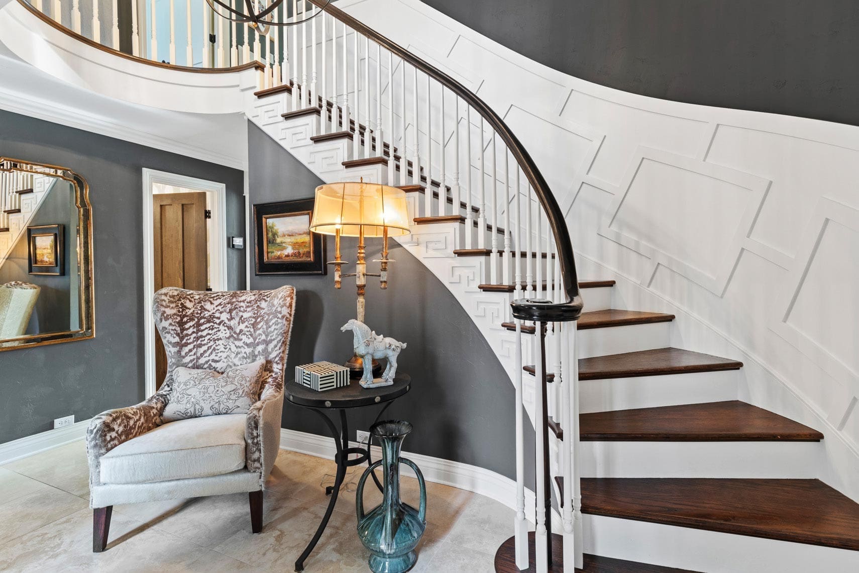 Residential real estate photo of a curved staircase with dark wood steps and white railing, featuring a patterned armchair and lamp beside a round table with decorative items, enhancing the homes interior design.