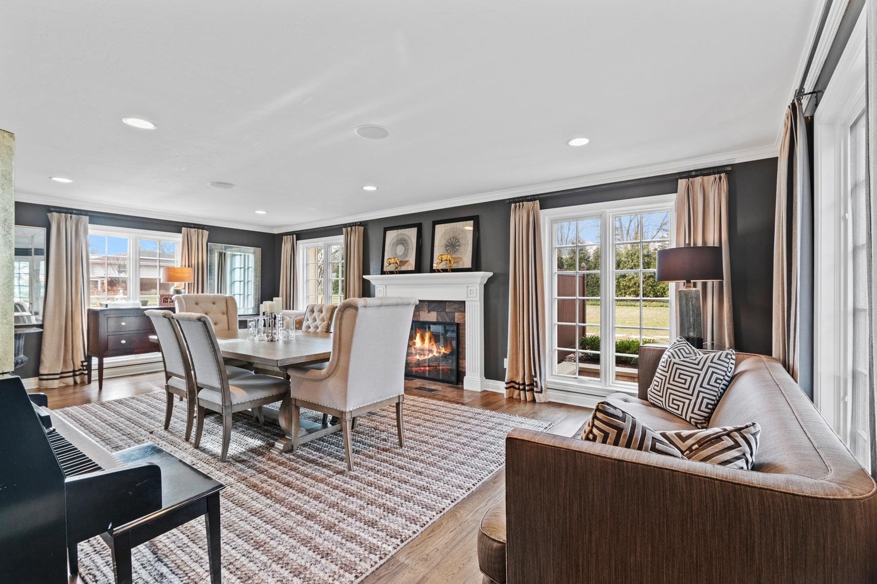 Spacious residential living room featuring a cozy fireplace, elegant dining area with table and chairs, comfortable sofa, and a piano. Large windows overlook the garden, while natural light illuminates the patterned rug on wooden flooring. Perfect for real estate photography showcasing inviting interiors.