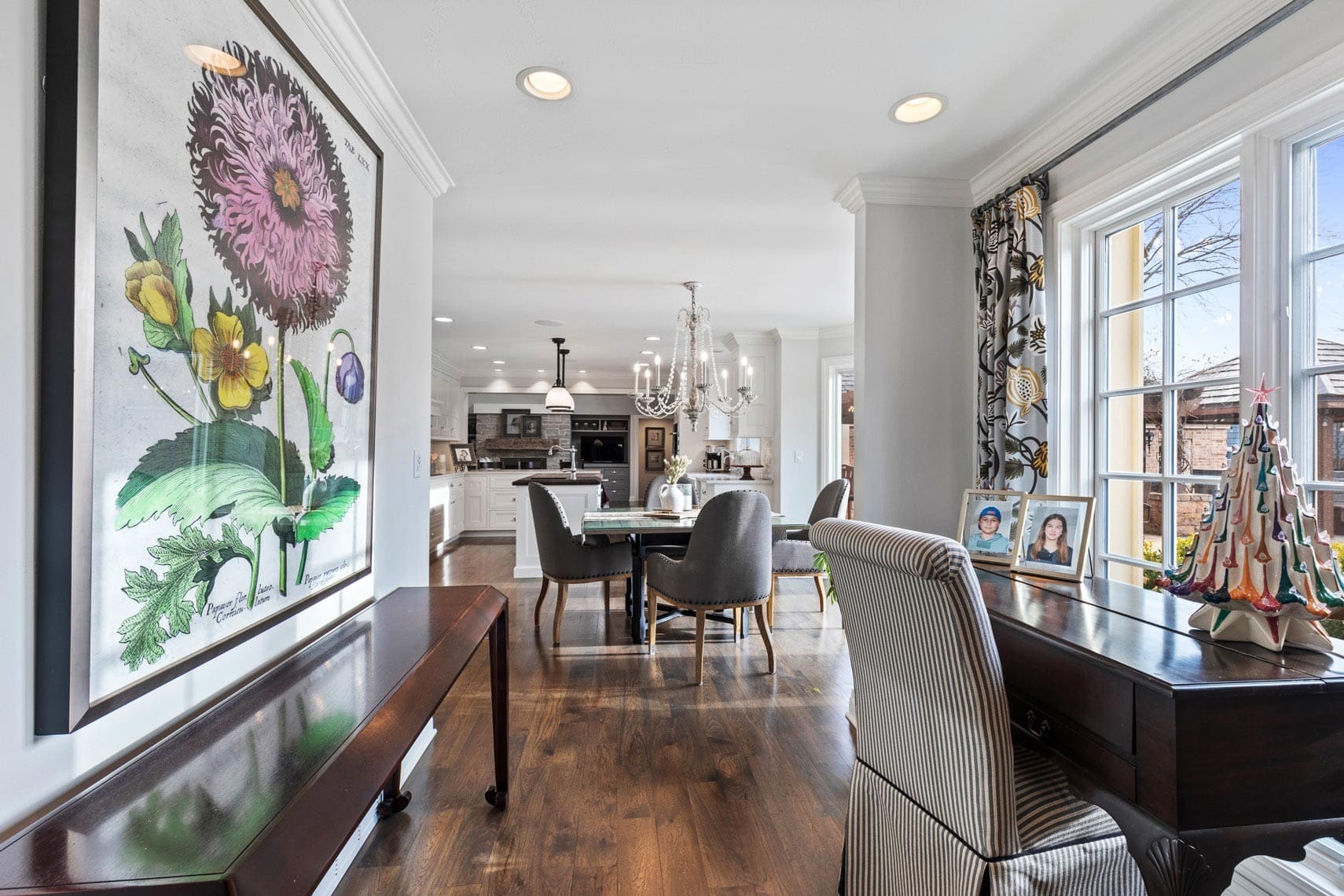 Spacious open-concept living room in a residential property featuring large botanical artwork, dark wooden dining table, striped chair, decorative tree, framed photos, and kitchen view. Perfect showcase for real estate photography.
