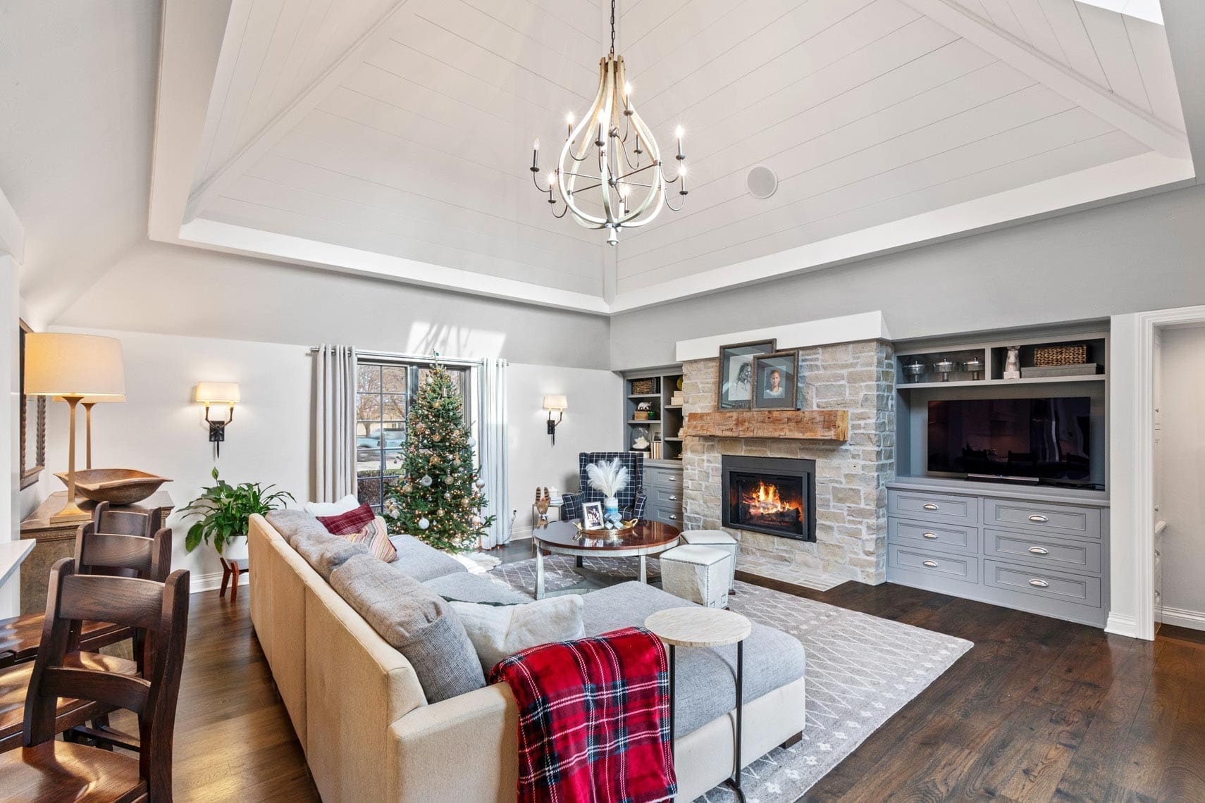 Residential real estate photo of a spacious living room featuring vaulted ceiling, chandelier, stone fireplace, Christmas tree, and beige sectional sofa with red plaid blanket; ideal for showcasing holiday-ready homes.
