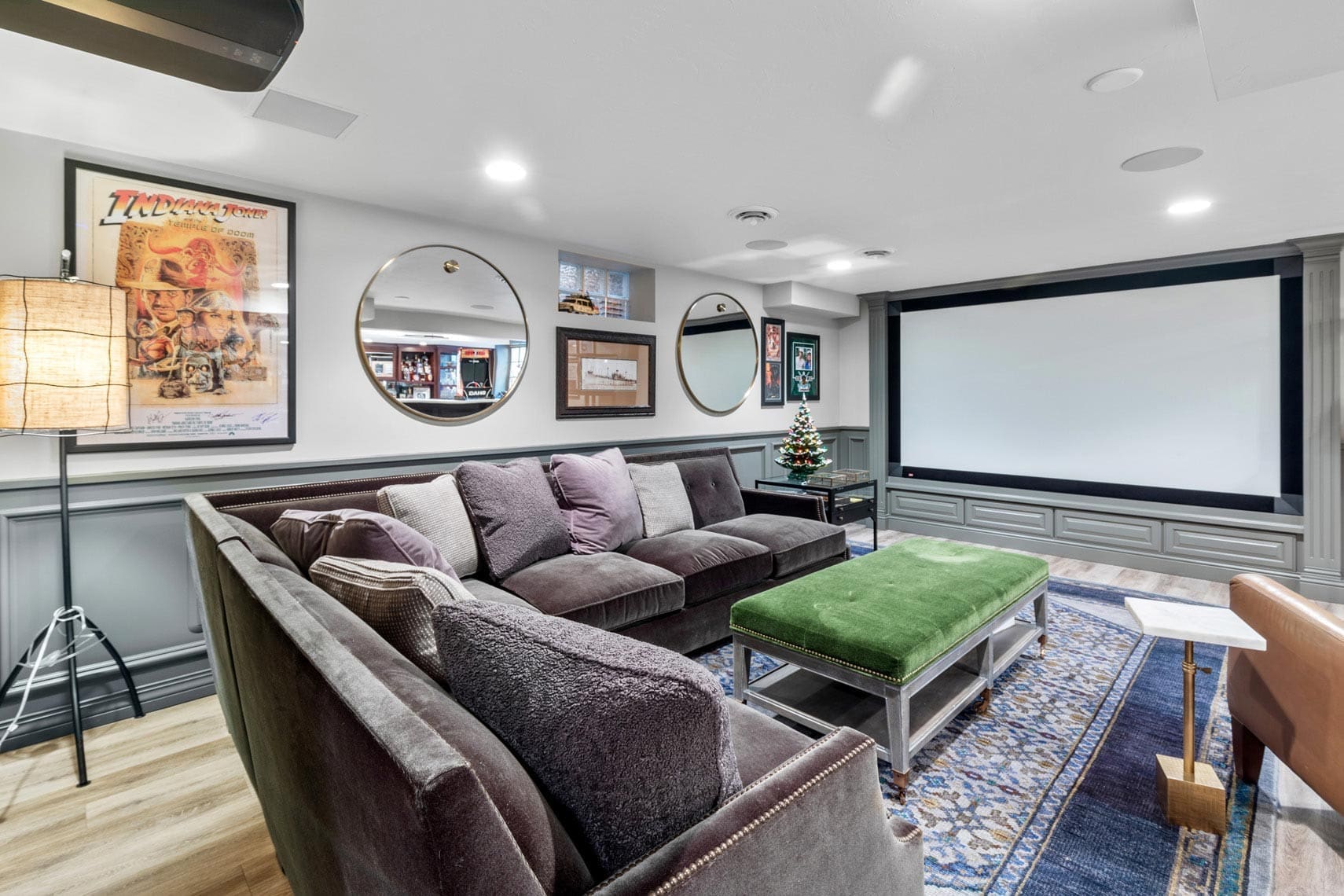 Real estate photography of a residential home theater room featuring a large projector screen, gray sectional sofa, green ottoman, and artistic decor including movie posters and round mirrors on the walls.