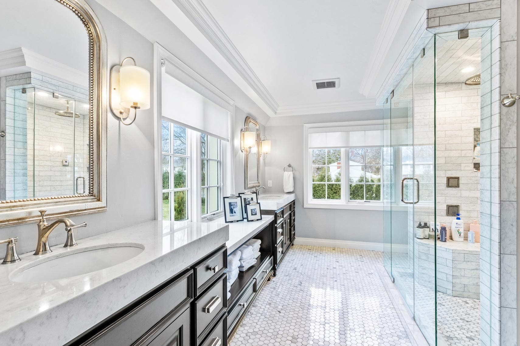 Luxury residential bathroom featuring dual sinks, large mirrors, and a glass-enclosed shower. Elegant white tiles, dark cabinets, and abundant natural light enhance the space from two windows.
