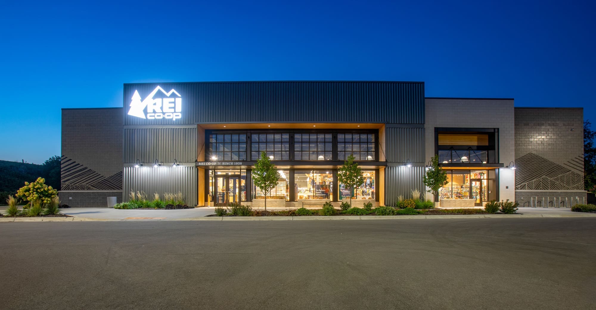 Commercial property for lease: REI Co-op retail storefront at dusk with bright signage, large glass windows, and minimal landscaping. Commercial property for lease: REI Co-op retail storefront at dusk with bright signage, large glass windows, and minimal landscaping.