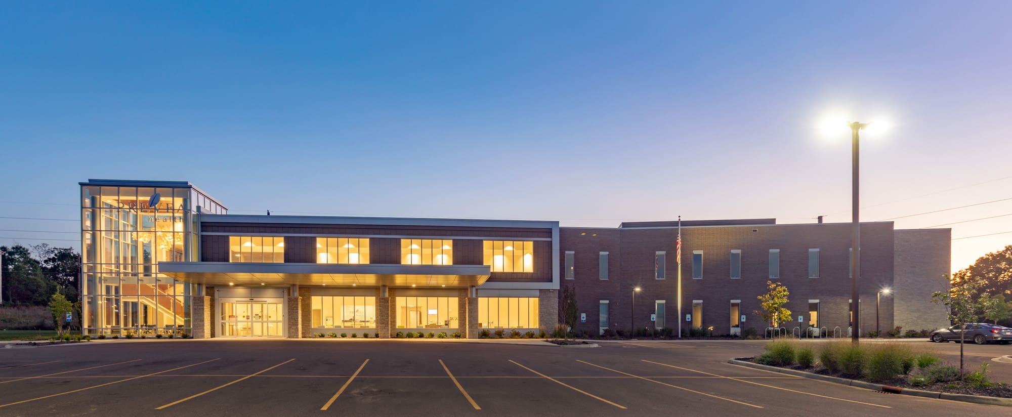 Commercial real estate photo of a modern two-story brick office building with large windows and glass stairwell at dusk, empty lot in front.