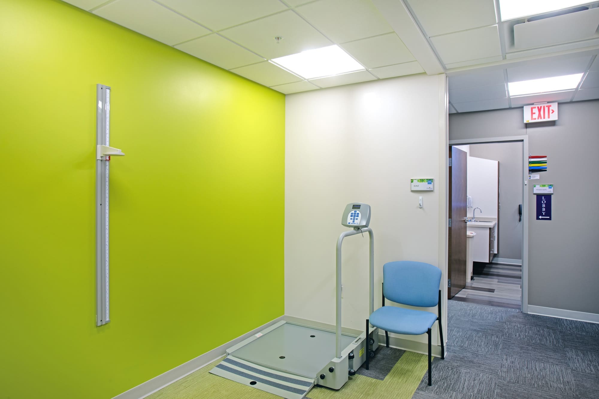 Commercial medical clinic interior featuring a green accent wall, modern equipment, blue chair, and hallway to sink area.