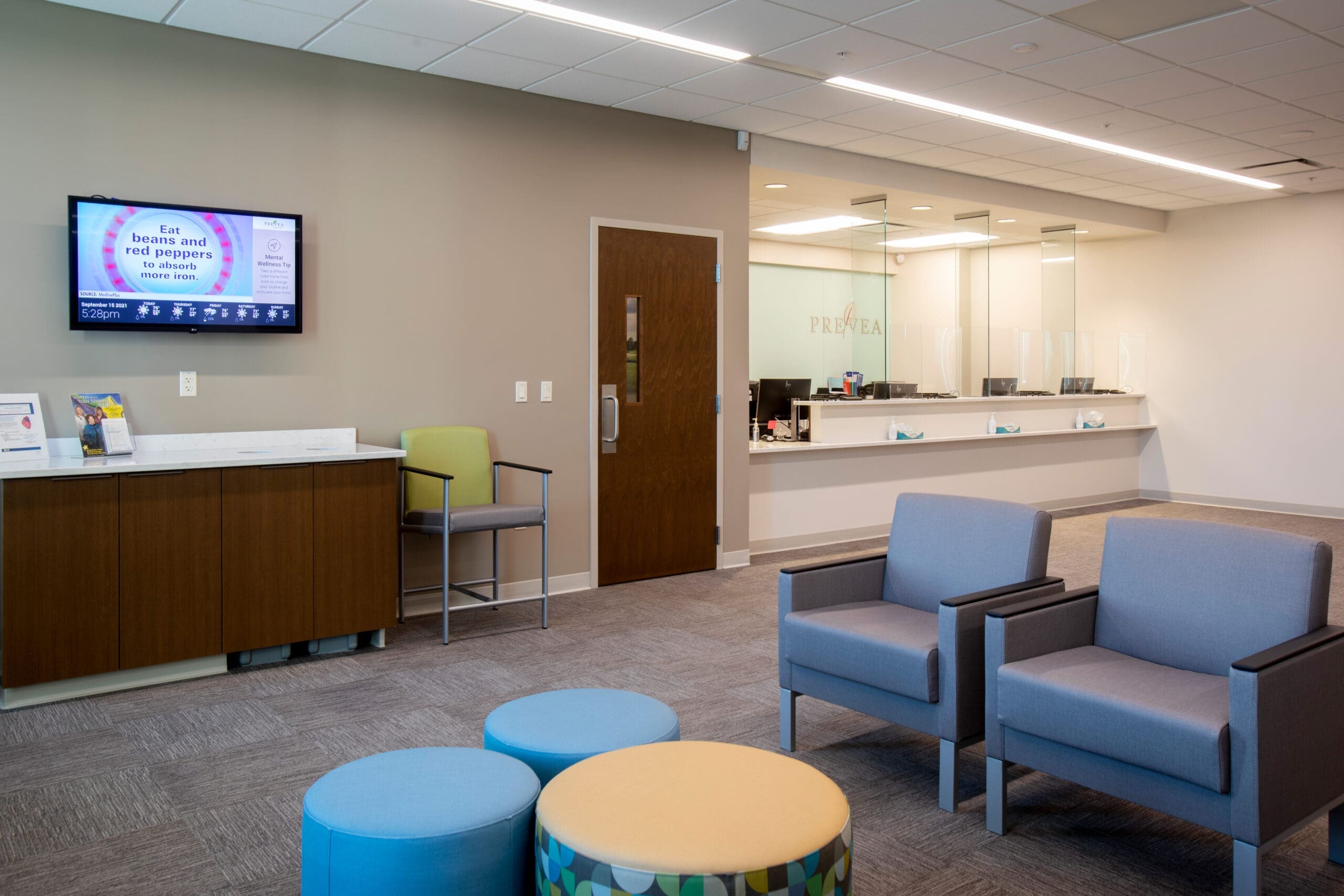 Commercial real estate clinic lobby with modern gray chairs, vibrant stools, TV, glass reception area, and bright lighting.
