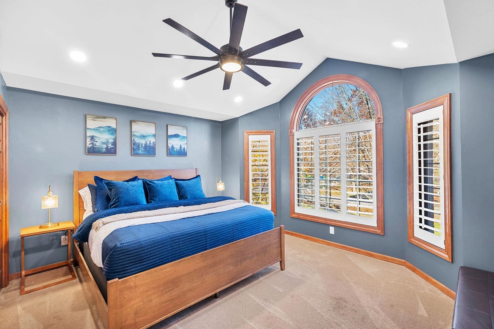 Residential bedroom showcasing a wooden bed with blue bedding, bedside tables featuring lamps, wall art, large window with shutters, and ceiling fan; ideal real estate photography for showcasing property interiors.