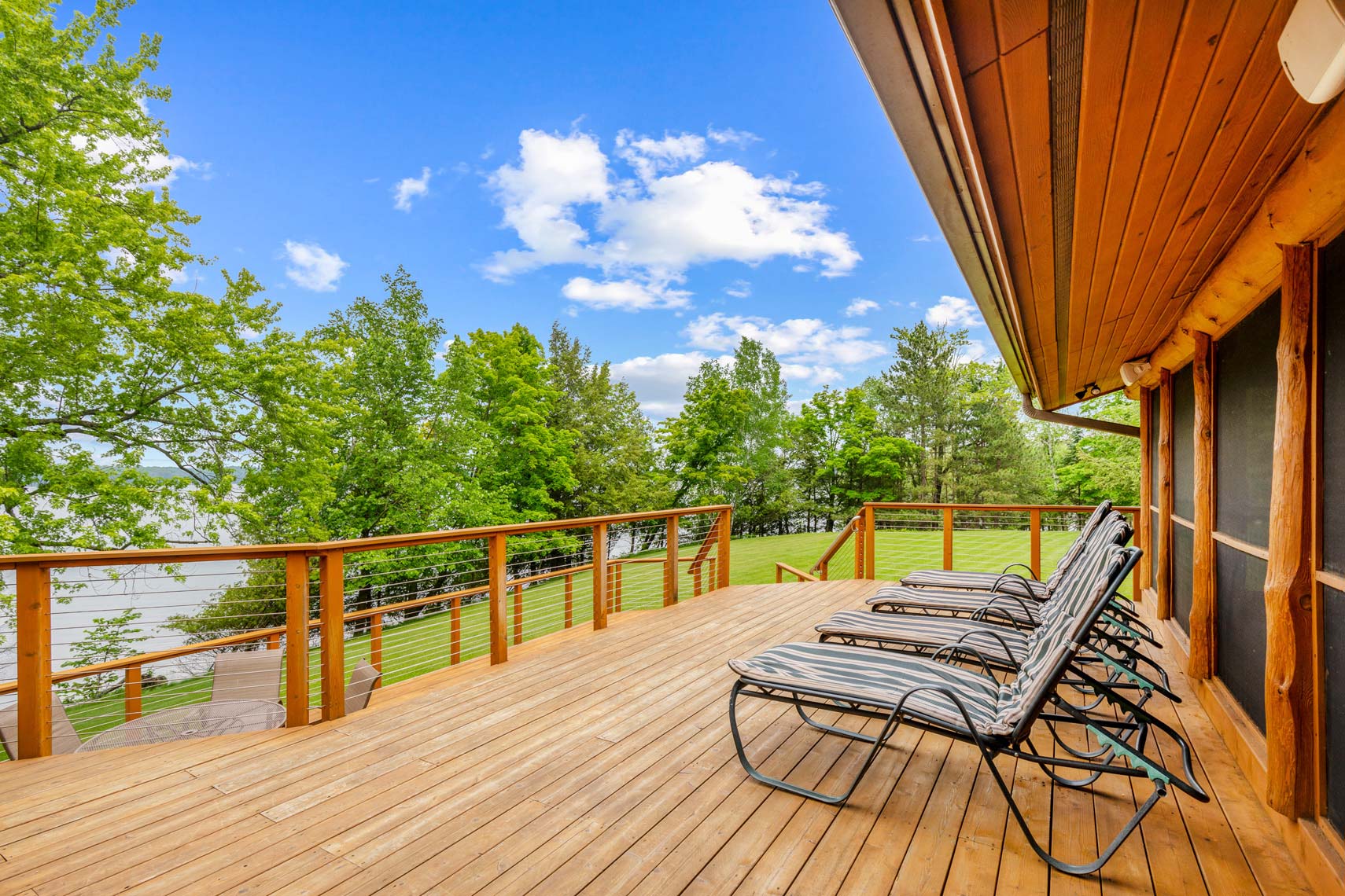 Real estate photography of a wooden deck with lounge chairs overlooking a scenic view of trees and a waterfront under a bright blue sky with scattered clouds, ideal for residential or vacation rental listings.
