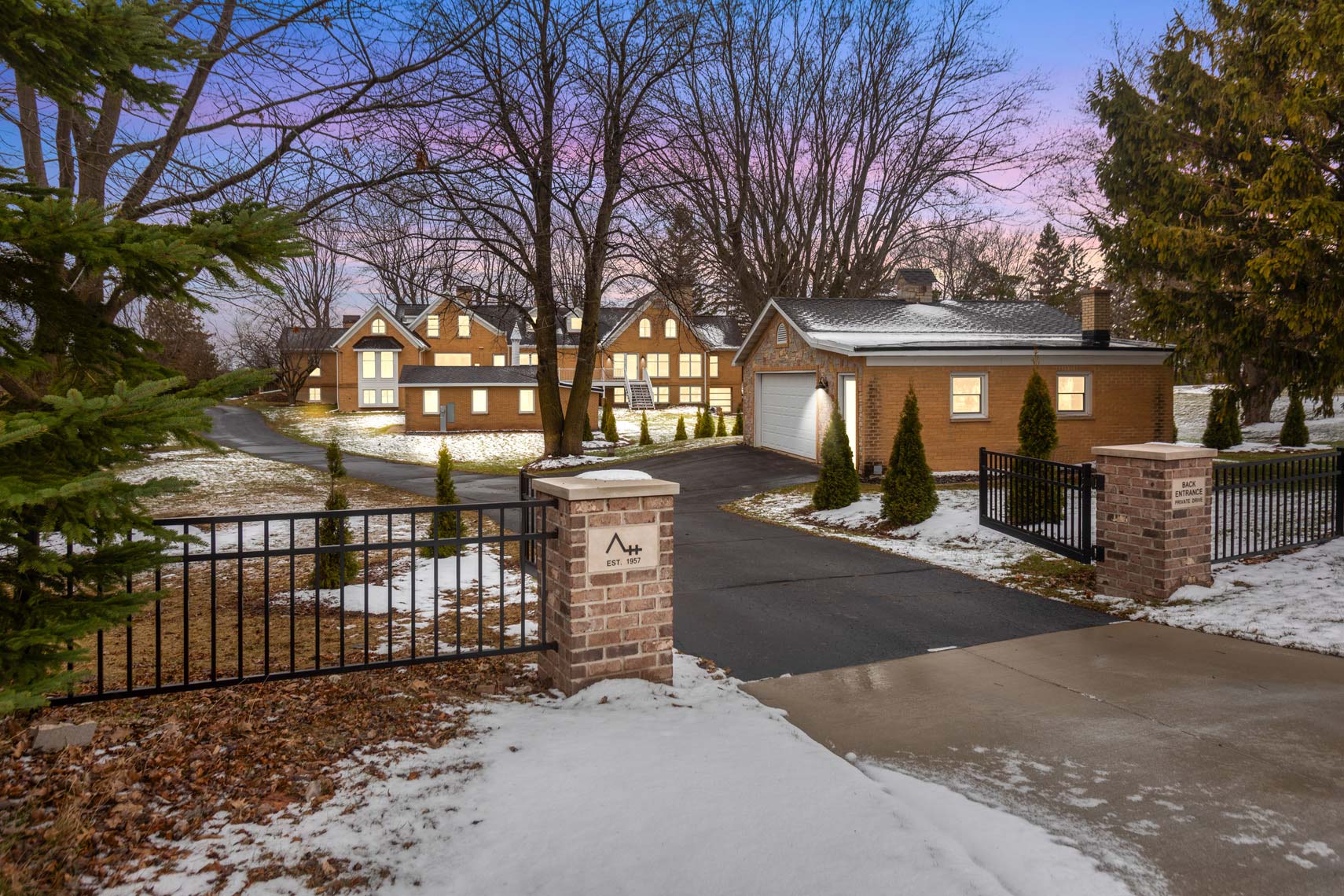 Commercial real estate photography of a spacious brick house featuring an attached garage, framed by a snowy landscape and mature trees, viewed through a black metal gate with brick posts at dusk.